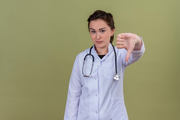 Smiling young doctor wearing medical gown wearing stethoscope her thumb down on green wall