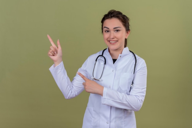 Smiling young doctor wearing medical gown wearing stethoscope points to side on green wall