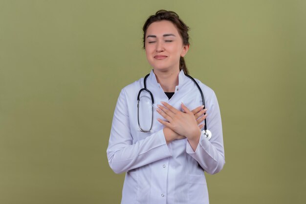 聴診器を身に着けている医療用ガウンを身に着けている笑顔の若い医者は緑の壁に彼女の手を心臓に置きます