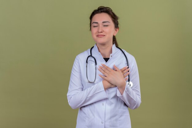 Smiling young doctor wearing medical gown wearing stethoscope put her hand on heart on green wall