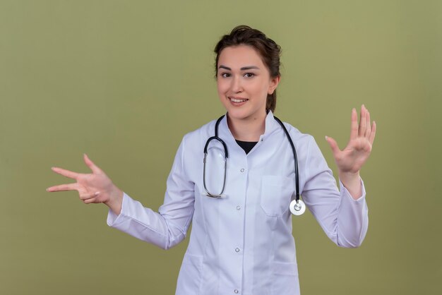 Smiling young doctor wearing medical gown wearing stethoscope shows different gesture on green wall