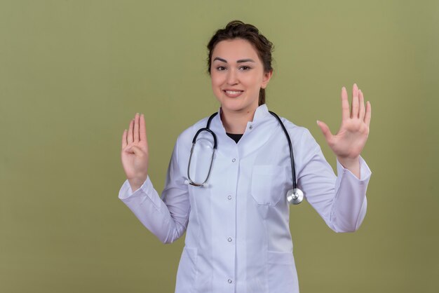 Smiling young doctor wearing medical gown wearing stethoscope shows different gesture on green wall