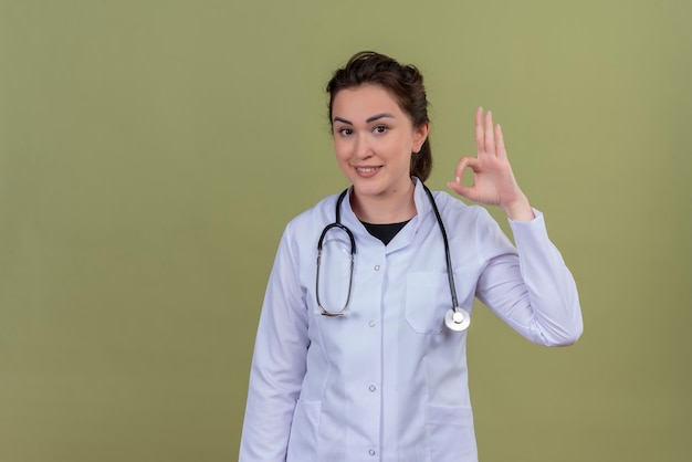 Улыбающийся молодой врач в медицинском халате со стетоскопом показывает жест на зеленой стене