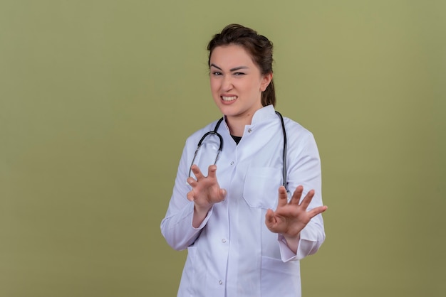 Smiling young doctor wearing medical gown wearing stethoscope shows tiger gesture on green wall