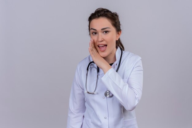 Smiling young doctor wearing medical gown wearing stethoscope whispers on white wall