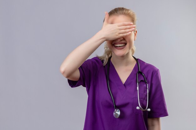 Smiling young doctor wearing purple medical gown and stethoscope covered eyes on isolated white wall