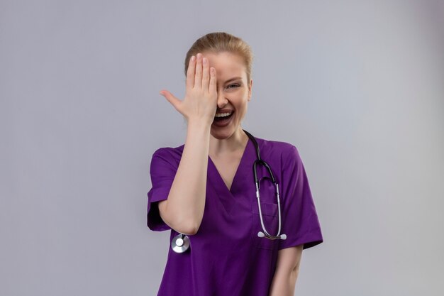 Smiling young doctor wearing purple medical gown and stethoscope covered one eye on isolated white wall