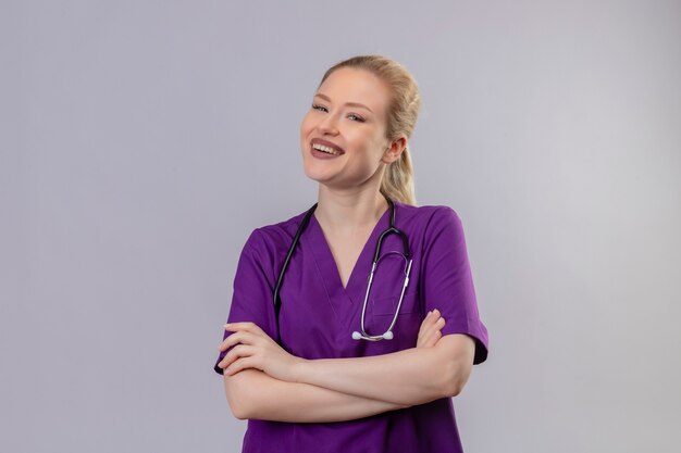 孤立した白い壁に紫色の医療用ガウンと聴診器の交差点の手を身に着けている若い医師の笑顔
