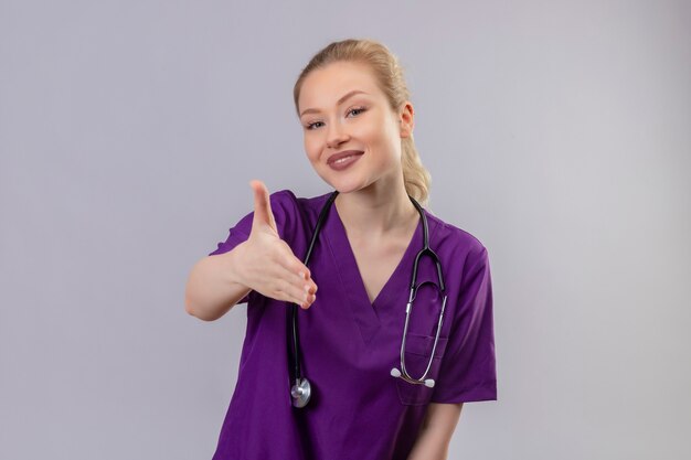 Smiling young doctor wearing purple medical gown and stethoscope held out hand on isolated white wall