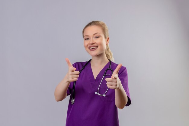Smiling young doctor wearing purple medical gown and stethoscope her thumbs up on isolated white wall