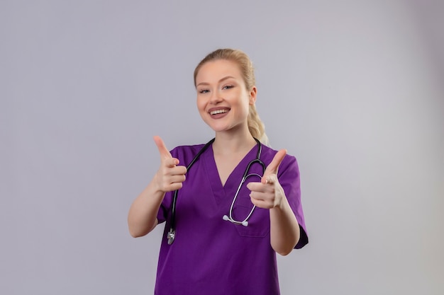 Улыбающийся молодой врач в фиолетовом медицинском халате и стетоскопе ее большие пальцы руки вверх на изолированной белой стене