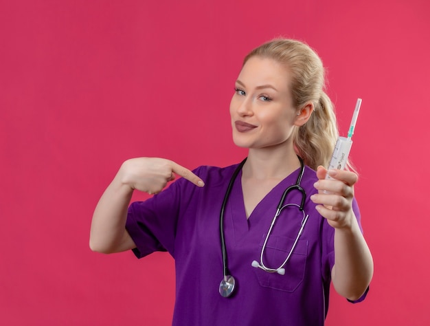 紫色の医療用ガウンと注射器を保持している聴診器を身に着けている笑顔の若い医者は、孤立したピンクの壁に自分自身を指しています