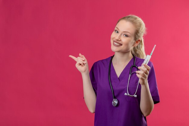 Il giovane medico sorridente che porta l'abito medico viola e lo stetoscopio che tiene la siringa punta a lato sulla parete rosa isolata