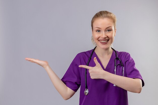 Smiling young doctor wearing purple medical gown and stethoscope points to side on isolated white wall