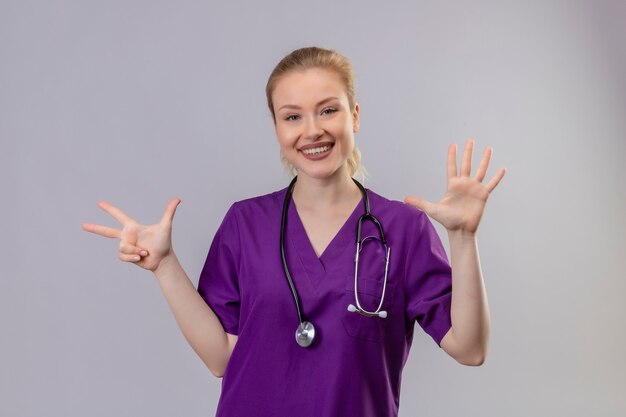 Улыбающийся молодой врач в фиолетовом медицинском халате и стетоскопе показывает другой жест на изолированной белой стене