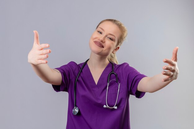 Smiling young doctor wearing purple medical gown and stethoscope shows hug gesture on isolated white wall