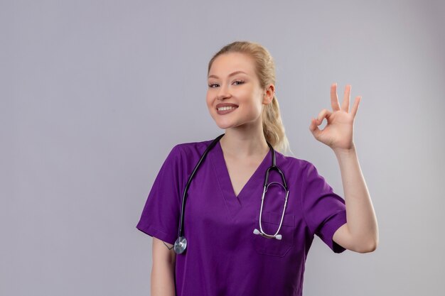 Smiling young doctor wearing purple medical gown and stethoscope shows okey gesture on isolated white wall