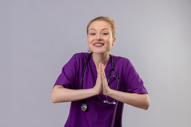 Il giovane medico sorridente che indossa l'abito medico viola e lo stetoscopio mostra pregare il gesto sulla parete bianca isolata