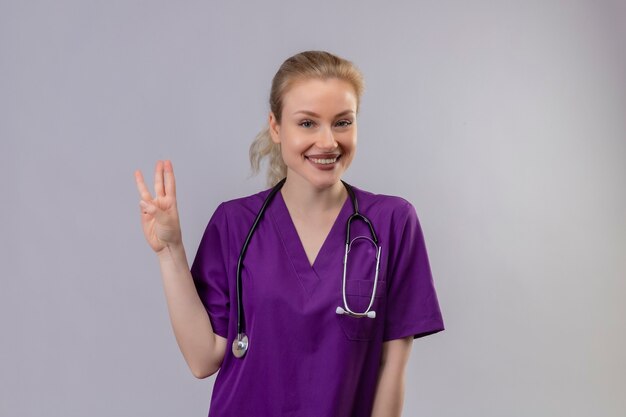 Smiling young doctor wearing purple medical gown and stethoscope shows three on isolated white wall