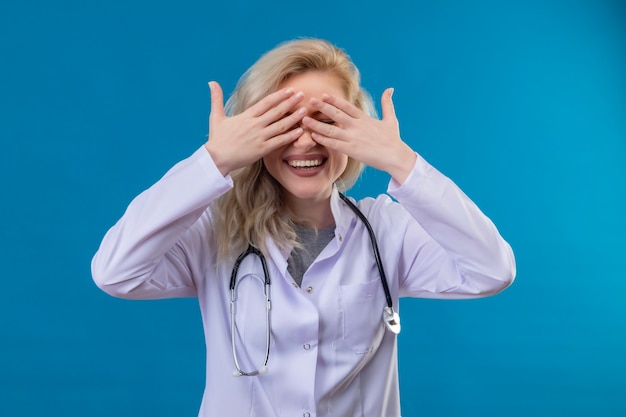 Бесплатное фото Улыбающийся молодой врач со стетоскопом в медицинском халате прикрыл глаза на синей стене