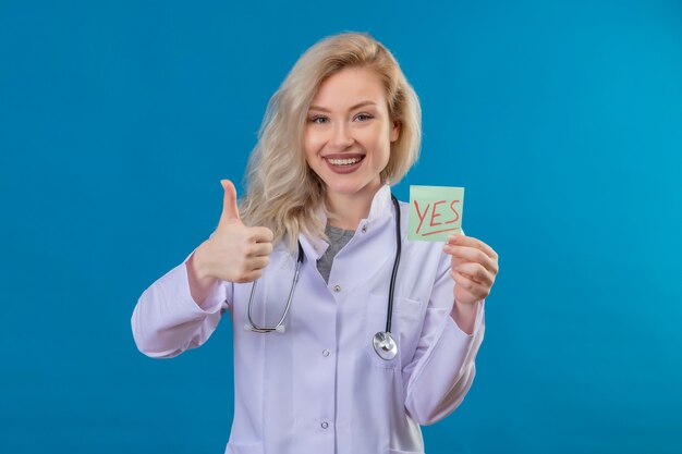 Smiling young doctor wearing stethoscope in medical gown holding paper yes mark her thumb up on blue wall