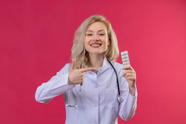 Smiling young doctor wearing stethoscope in medical gown holding pills and points on red backgroung