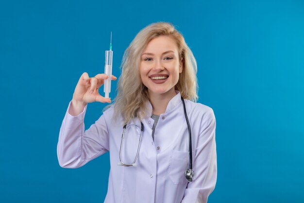 青い壁に注射器を保持している医療用ガウンで聴診器を身に着けている若い医者の笑顔