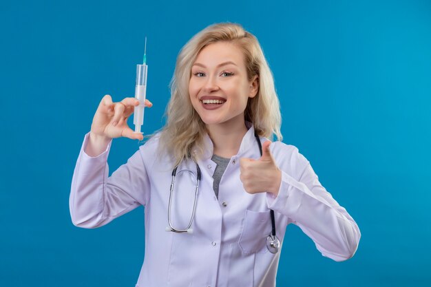Smiling young doctor wearing stethoscope in medical gown holding syringe her thumb up on blue wall
