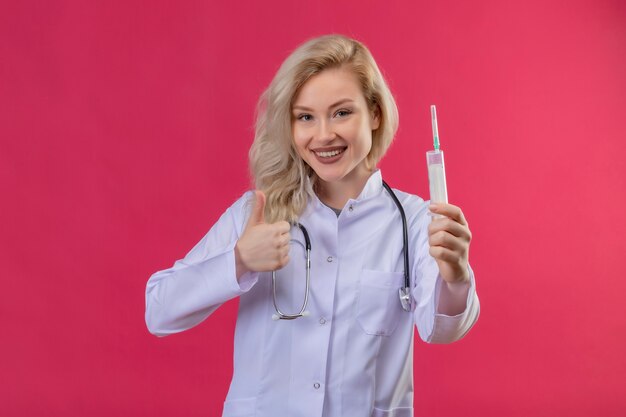 赤い背景に彼女の親指を上に注射器を保持している医療用ガウンで聴診器を身に着けている若い医者の笑顔
