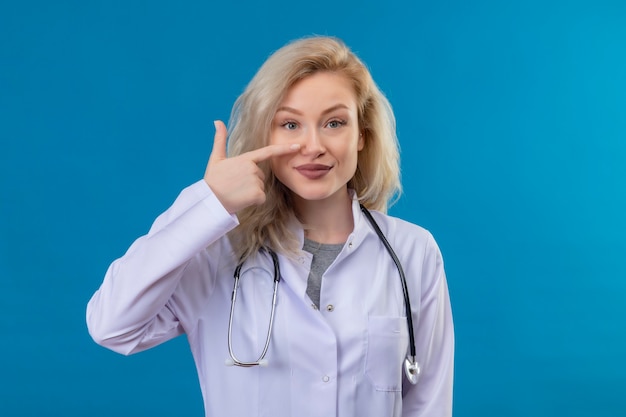 Smiling young doctor wearing stethoscope in medical gown points to nose on blue wall