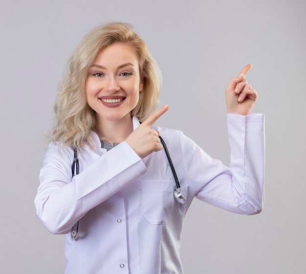 Smiling young doctor wearing stethoscope in medical gown points to side on white wall