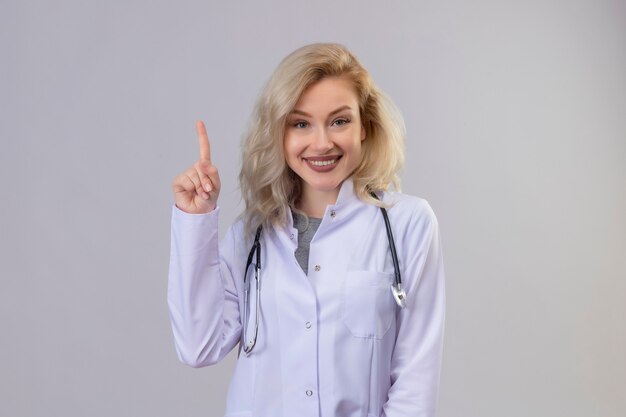 Smiling young doctor wearing stethoscope in medical gown points to up on white wall