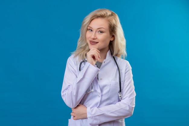 Free photo smiling young doctor wearing stethoscope in medical gown put her hand under jaw on blue wall