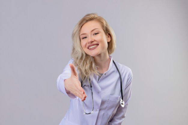 白い壁に挨拶のジェスチャーを示す医療用ガウンで聴診器を身に着けている若い医師の笑顔