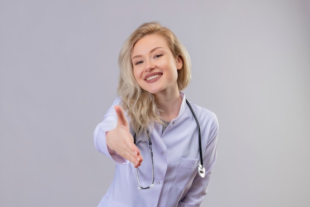 Smiling young doctor wearing stethoscope in medical gown showing greeting gesture on white wall