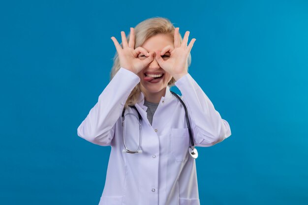 Smiling young doctor wearing stethoscope in medical gown showing looking gesture on blue wall