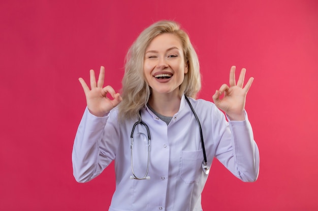 Free photo smiling young doctor wearing stethoscope in medical gown showing okey gesture both hands on red backgroung