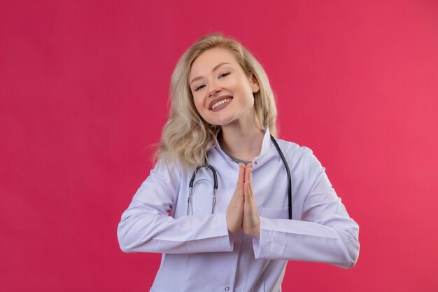 Smiling young doctor wearing stethoscope in medical gown showing pray gesture on red backgroung