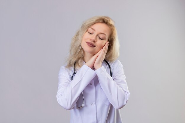 Smiling young doctor wearing stethoscope in medical gown showing sleep gesture on white wall