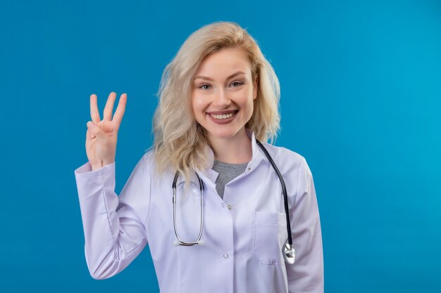 파란색 벽에 3을 보여주는 의료 가운에 청진기를 입고 웃는 젊은 의사