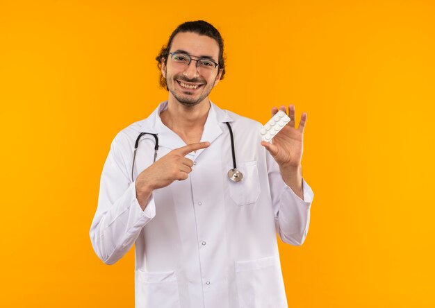 Smiling young doctor with medical glasses wearing medical robe with stethoscope holding and points to pills on isolated yellow wall with copy space