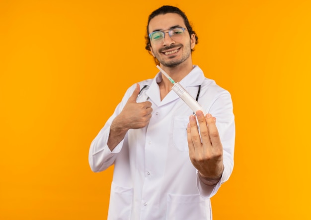 Smiling young doctor with medical glasses wearing medical robe with stethoscope holding syringe his thumb up on isolated yellow wall with copy space