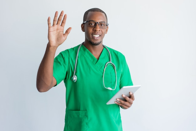 Smiling young doctor with touchpad waving hand.