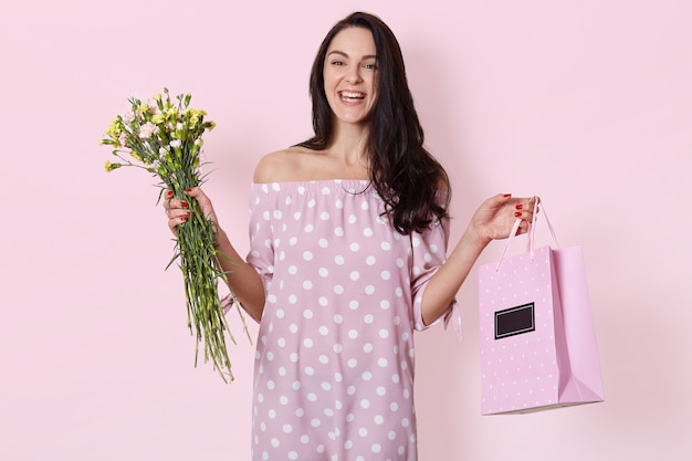 smiling young European woman has dark long wavy hair, wearing polka dot rose dress, holds gift bag and bouquet of flowers, posing on light pink, has birthday.