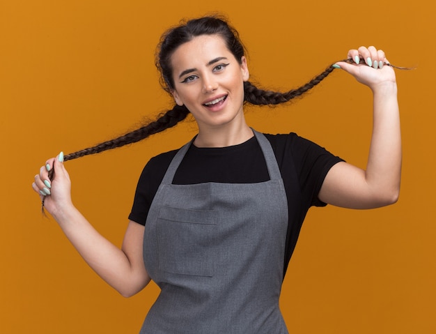 Smiling young female barber in uniform grabbed hair isolated on orange wall