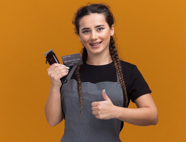 Smiling young female barber in uniform holding credit card and hair clippers showing thumb up isolated on orange wall