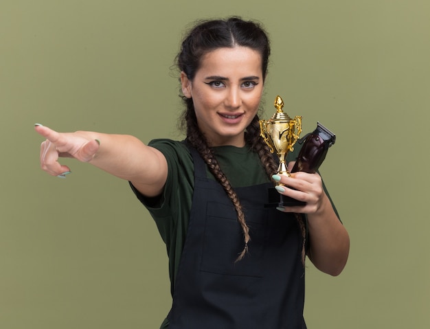 Smiling young female barber in uniform holding hair clippers with winner cup points at side isolated on olive green wall
