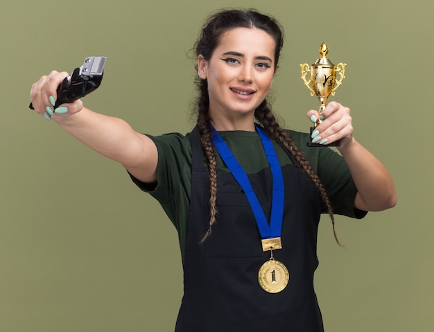 Smiling young female barber in uniform and medal holding winner cup and holding out hair clippers at camera isolated on olive green wall