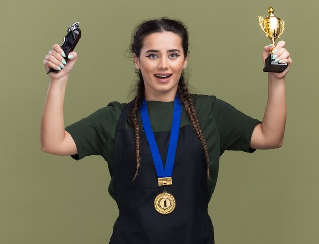 オリーブグリーンの壁に隔離されたバリカンと制服とメダルを上げる勝者カップで若い女性の理髪師の笑顔