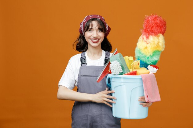 Smiling young female cleaner wearing uniform and bandana holding bucket of cleaning tools with both hands looking at camera isolated on orange background